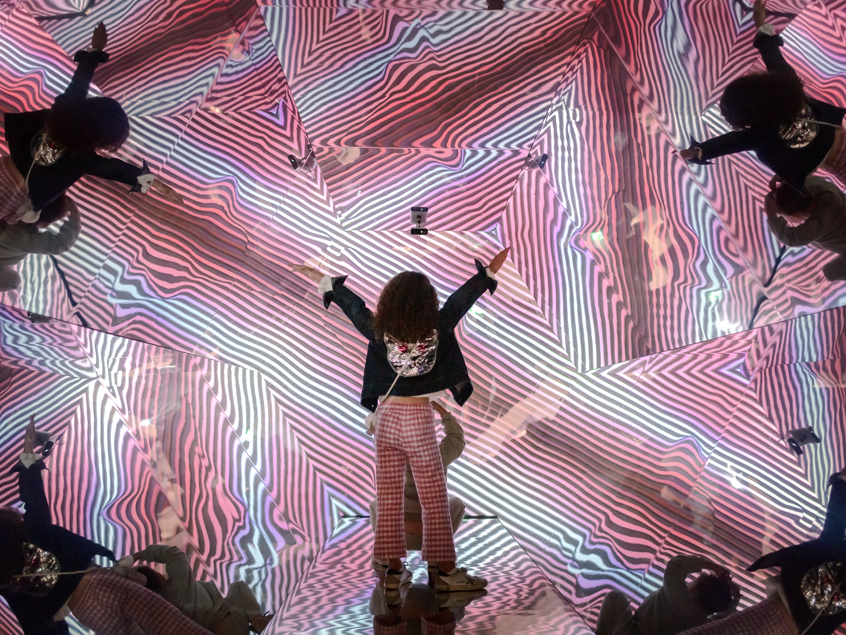 A child raises both arms in the Kaleidoscope Room, reflected many times among pink and white stripes