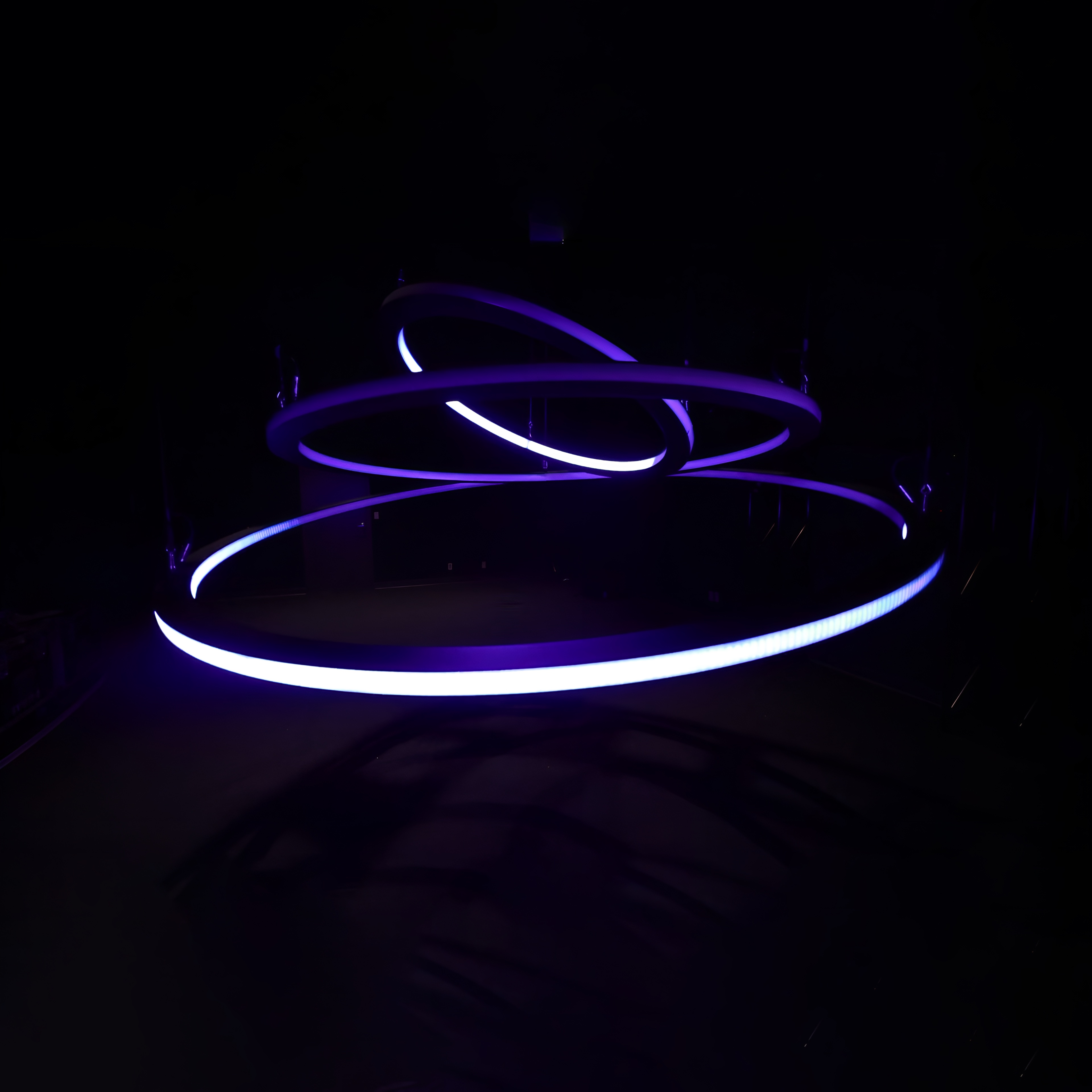 Kinetic chandelier of three violet-lit LED rings, tilted at different angles in a dark room