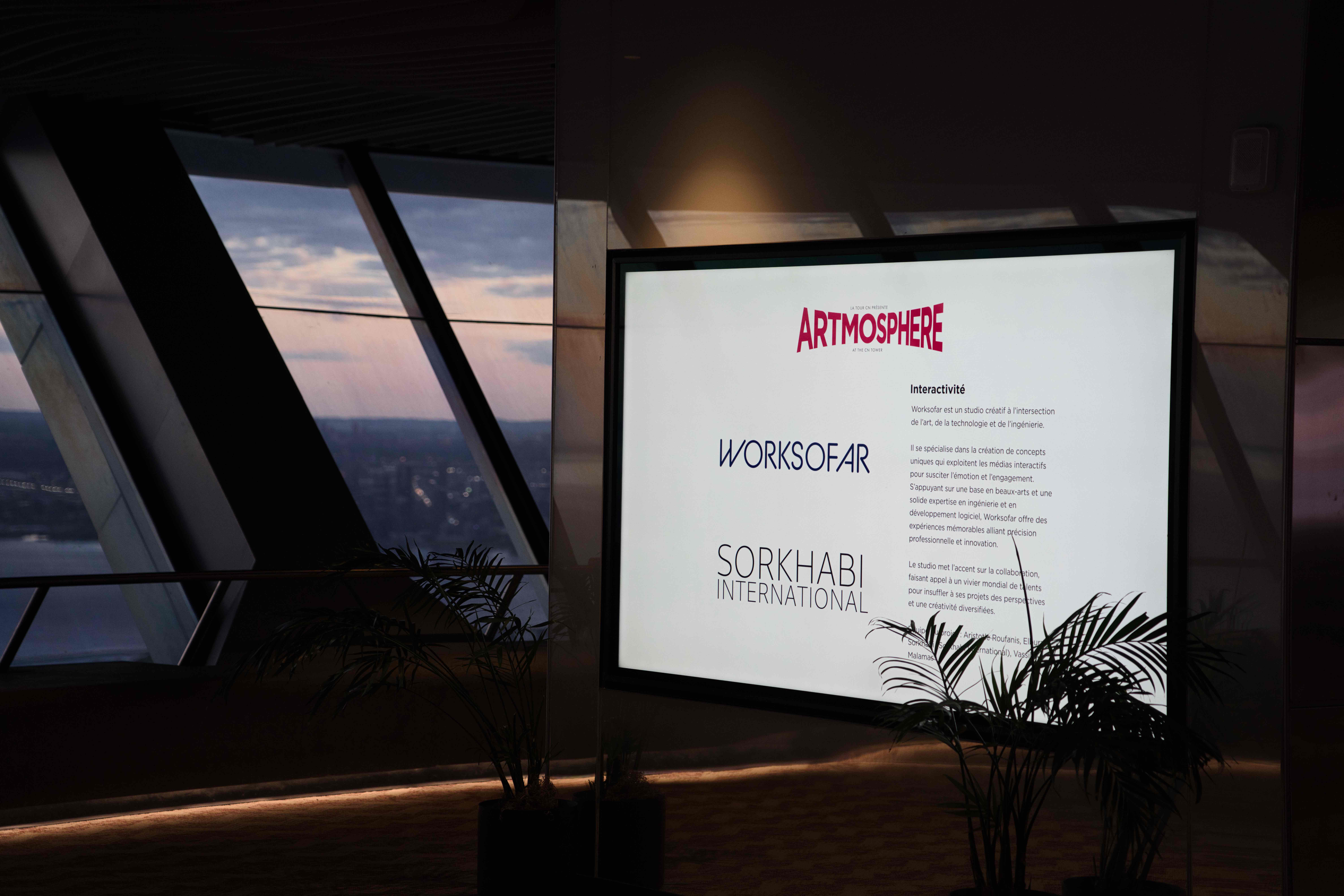 Screen crediting Artmosphere, Worksofar and Sorkhabi International beside windows overlooking the city at dusk