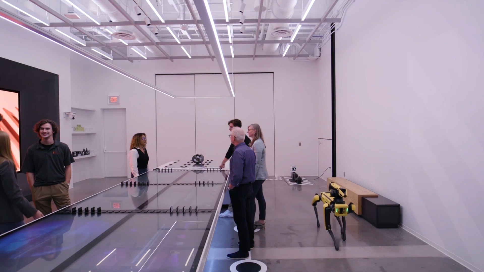 Visitors along a long display table in a bright white space at The Smart Factory, with a four-legged robot