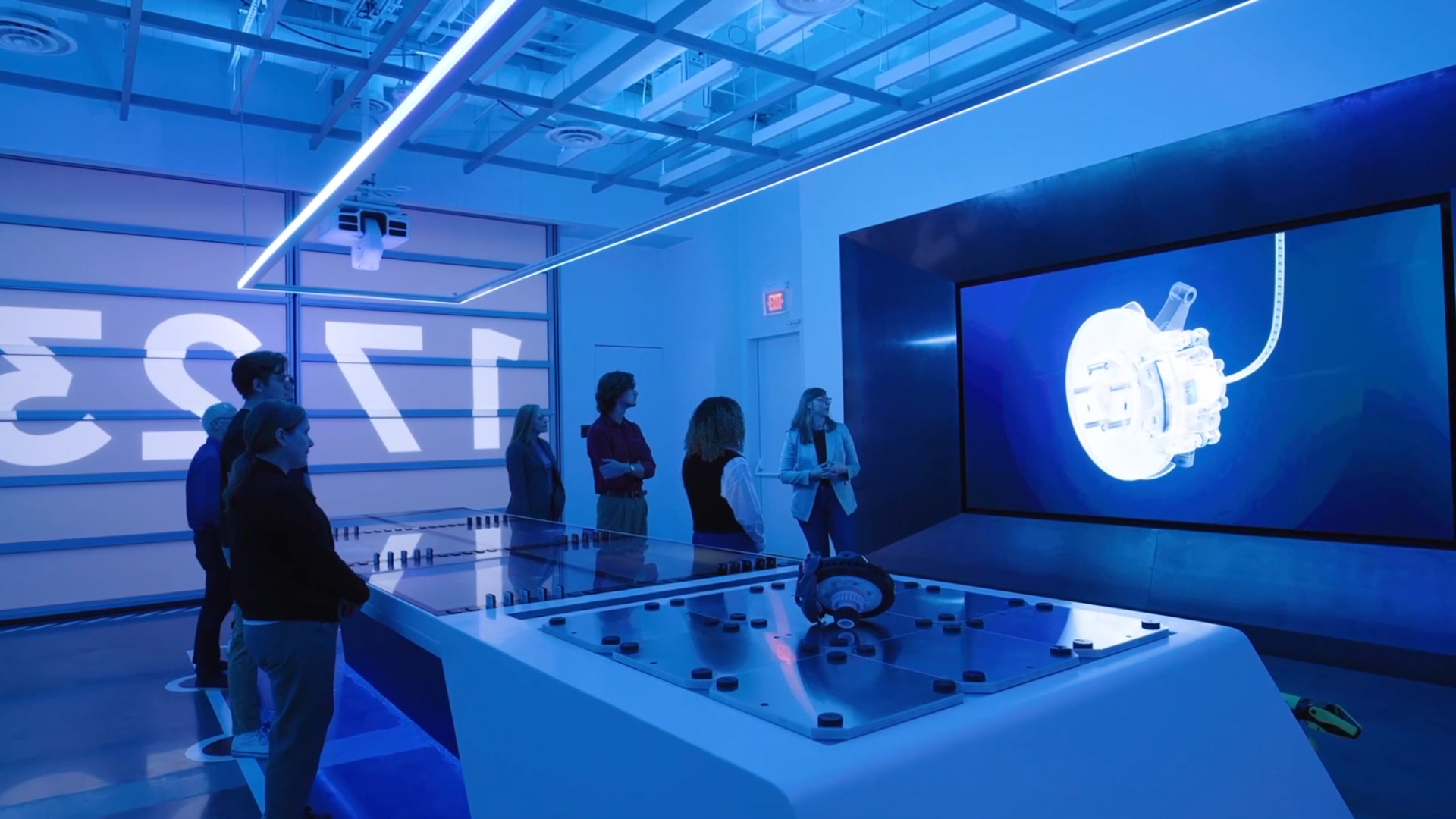 Visitors in a blue-lit room at The Smart Factory, beside a large screen showing a 3D machine part