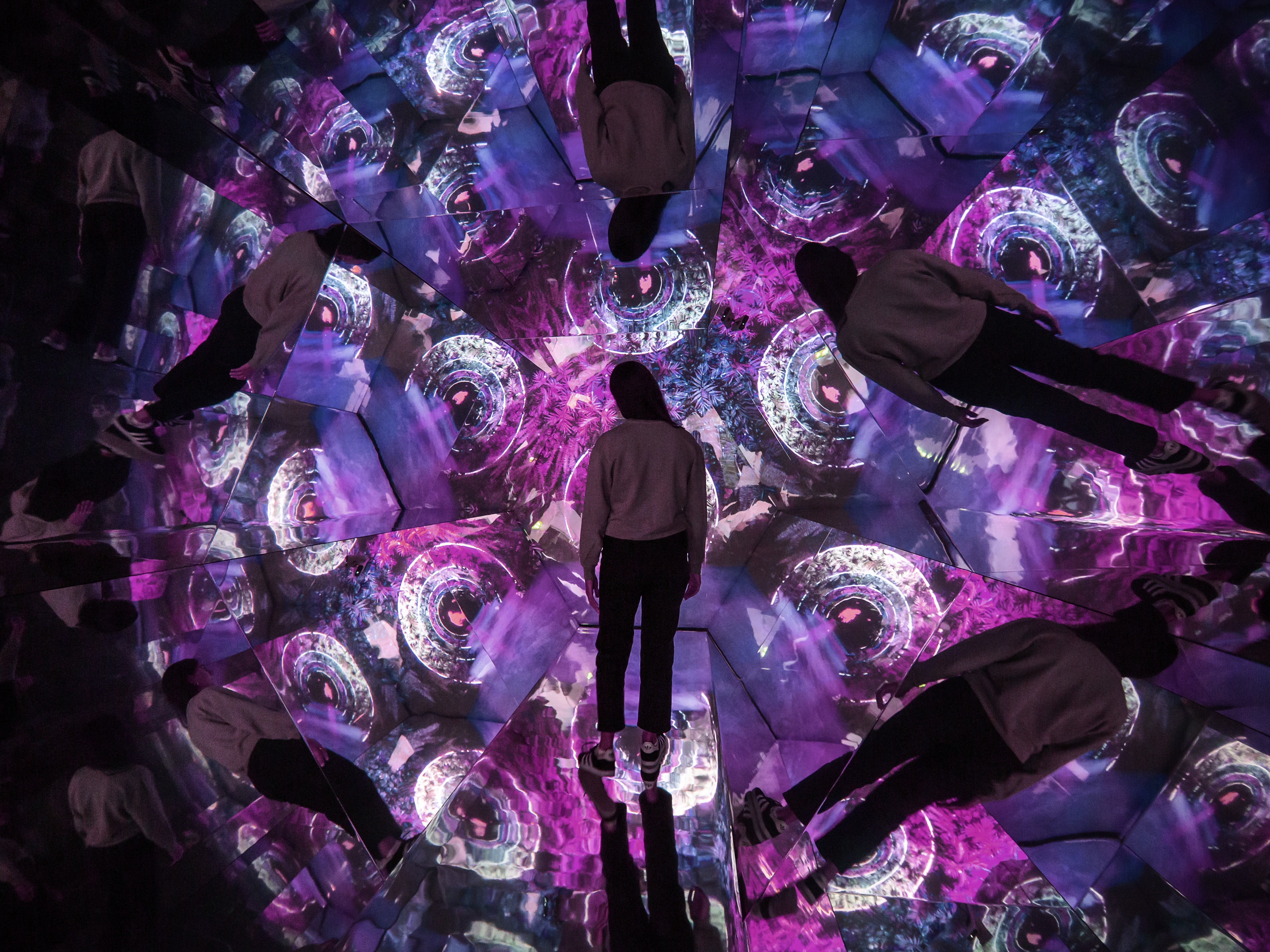 A visitor stands in the Kaleidoscope Room, surrounded by mirrored reflections of glowing purple patterns